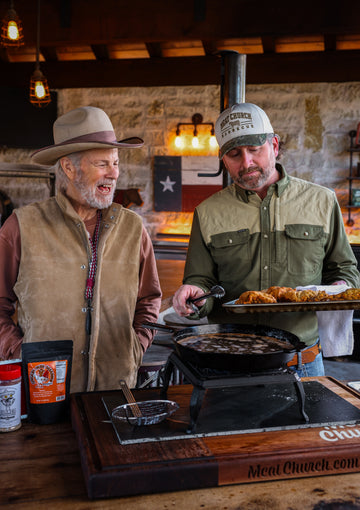 Two men prepare food beside a grill in a rustic kitchen, with a tray of cooked wings and sauces