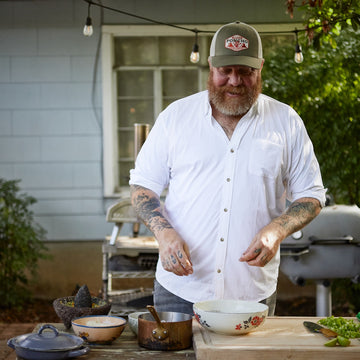 Bearded man in a white button-down shirt preparing food outdoors beside a barbecue grill and vegetables