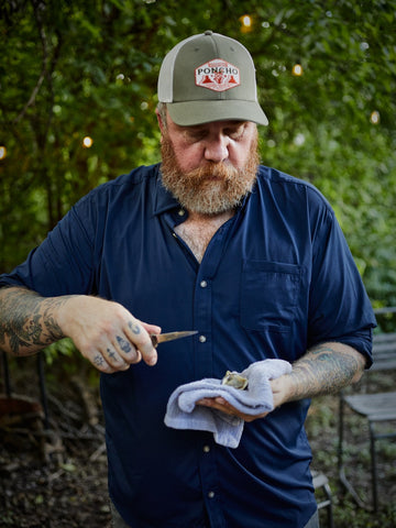 Bearded man in a navy shirt and Poncho Outdoors cap wipes a small tool with a cloth outdoors