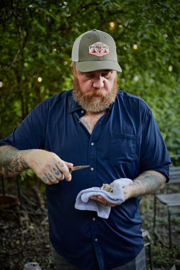 Bearded man in a navy shirt and Poncho Outdoors cap wipes a small tool with a cloth outdoors