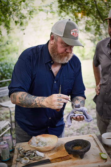Bearded man in a navy shirt shucks oysters at an outdoor table beside another person