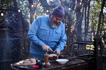 Bearded man in a blue shirt slicing grilled meat at an outdoor barbecue surrounded by trees and string lights