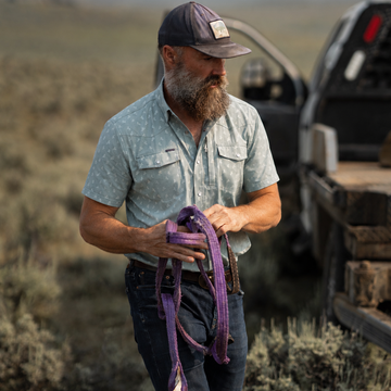 Man outdoors wearing The Lupine shirt holding purple straps near truck in rugged landscape
