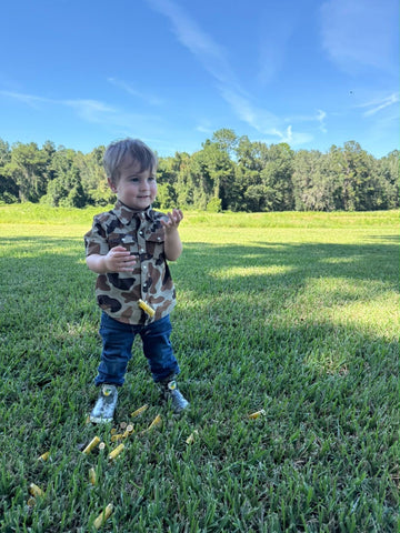 Young child in a camouflage shirt and jeans standing in a sunny grassy field with trees behind
