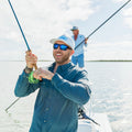 man reels in fly while wearing a deep blue long sleeve fishing shirt