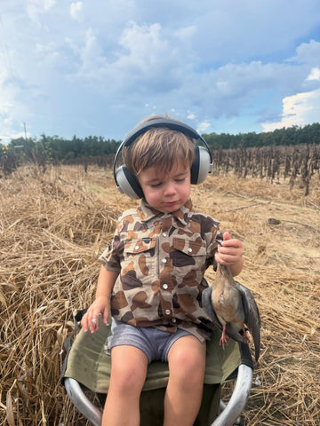 Child wearing headphones sits in a field holding a bird and wearing a camouflage button shirt