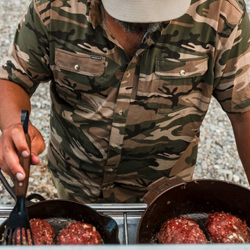 Man in camo shirt grilling burgers outdoors featuring The Asado shirt