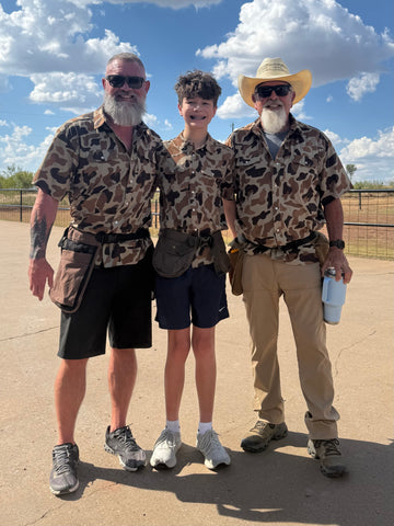Two men and a teenager wearing camouflage outdoor shirts pose beneath a bright blue sky.
