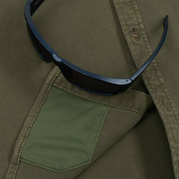 Close-up of an olive green shirt pocket with a pair of black sunglasses resting on The Buck Snort shirt outdoorswear.