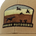 Close-up of a brown fabric patch on a hat featuring a dog, flying birds, and the text "Poncho Outdoors" in embroidered letters.