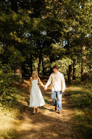 Man in a light shirt and jeans walks hand in hand with a woman in a white dress along a wooded trail