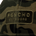 Close-up of a camouflage fabric patch with the embroidered text "PONCHO OUTDOORS" in green and beige tones.