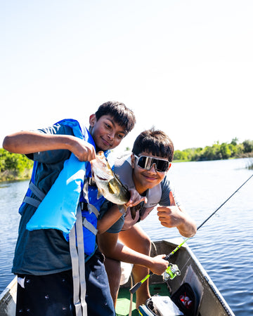 Two boys on a boat celebrate a fishing catch, with one holding a fish and the other giving a thumbs up