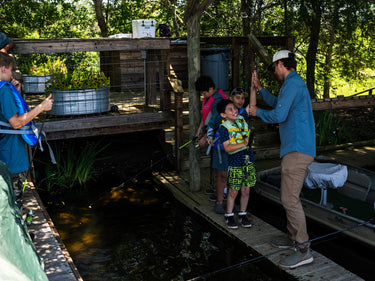 People and children gather at a shaded fishing pond beneath a mounted fish display
