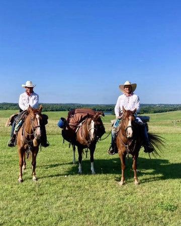 Two horseback riders in cowboy hats alongside a saddled pack horse in a sunny green field