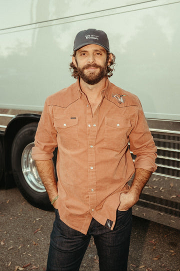 Bearded man wearing a rust-colored button-up shirt and black cap beside a vintage truck