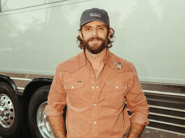Bearded man wearing a rust colored button down shirt and cap beside a large trailer