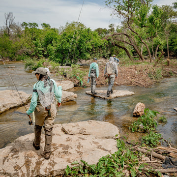 Three anglers in outdoor shirts wade through a shallow rocky stream surrounded by trees