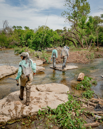 Anglers wading through a shallow forest stream with fishing rods and outdoor shirts