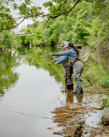 Two anglers in long-sleeved shirts fishing beside a calm, tree-lined river with a landing net