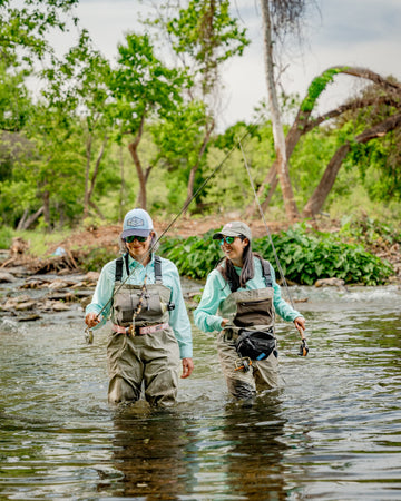 Two anglers in waders and fishing shirts standing in a shallow forest stream with rods in hand