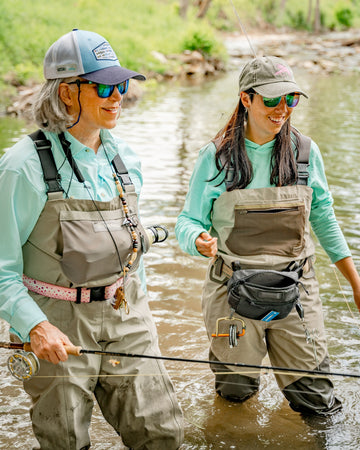 Two women wearing waders and shirts while fly fishing in a shallow stream