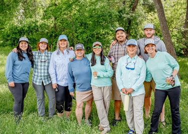 Group of nine people wearing outdoor shirts and hats, standing together in a lush green forest setting