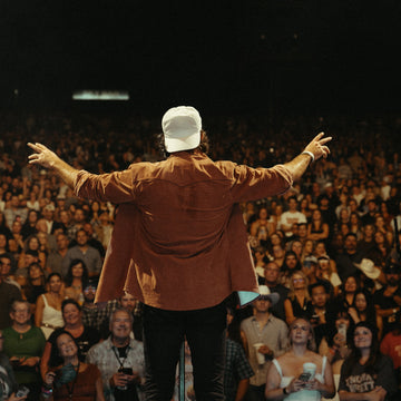 Performer in a brown shirt and white cap extends his arms toward a cheering crowd