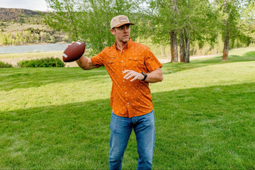 Man holding a football on a grassy lawn near a lake, wearing an orange patterned button-up shirt