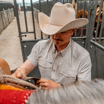 Man putting saddle on a horse wearing short sleeve pearl snap