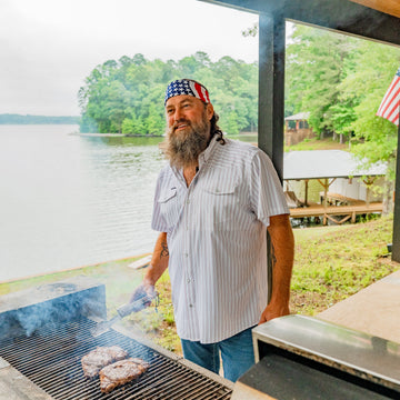 Bearded man in a striped shirt grilling burgers beside a lakeside shelter with trees and an American flag