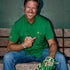 Man in a green button-up shirt and jeans sitting on a wooden bench holding a baseball and green glove, smiling and pointing at his shirt logo.