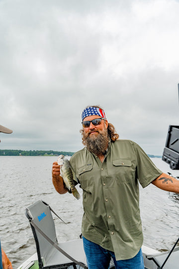Bearded man in a green shirt holds a fish aboard a boat on a lake under cloudy skies