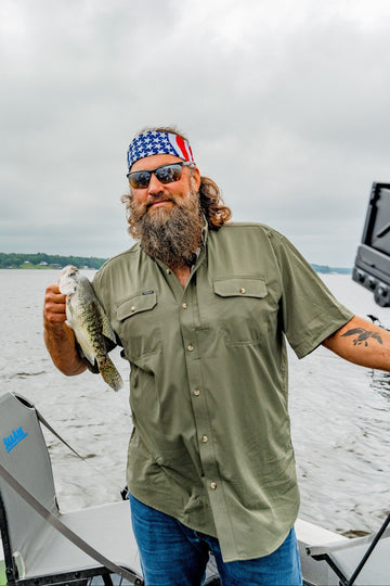 Bearded man in sunglasses and a bandana holding a fish aboard a boat on a cloudy lake