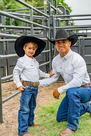 Adult and child wearing cowboy hats and white shirts beside metal livestock pens outdoors