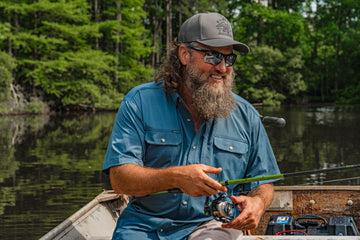 Bearded man in a blue shirt and cap fishing from a boat on a wooded lake