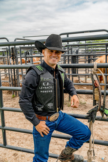 Man in a black cowboy hat and Poncho shirt standing beside a livestock pen with ropes and boots