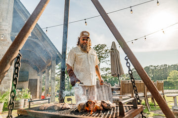Bearded man in a patterned shirt grilling meat on an outdoor barbecue beside a wooden deck