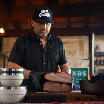 Man in a black shirt and cap prepares sliced meat on a cutting board in a rustic kitchen
