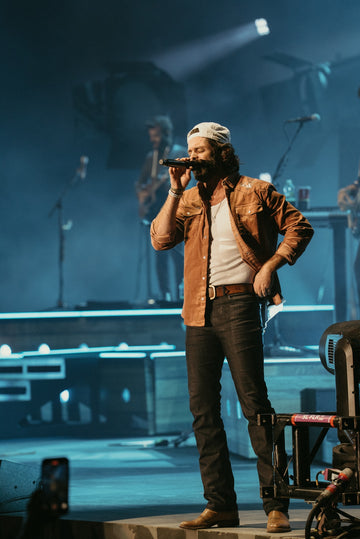 Singer in a brown jacket and knit cap performing into a microphone on a blue-lit stage