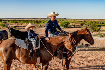 A cowboy and child wearing hats ride horses across a sunlit desert landscape