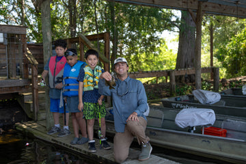 An adult and three children pose on a wooden dock beside a canoe in a wooded outdoor setting