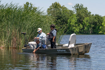 Two people in a small boat on a lake beside tall reeds and trees in the outdoors