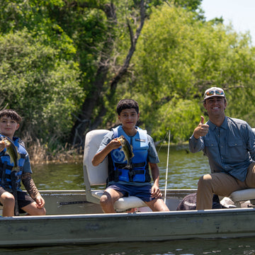 Three people in a boat on a tree-lined lake, with two children wearing blue life jackets and an adult in a blue shirt