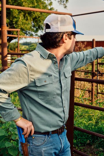 Man wearing a green button-down shirt and mesh-back cap leaning against a ranch fence outdoors