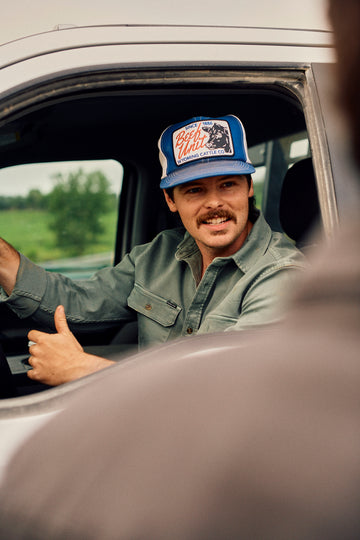 Man in a vehicle wearing a blue graphic cap and green button up shirt, giving a thumbs up