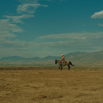 Horseback rider crossing a wide dry plain with rugged mountains beneath a cloudy blue sky