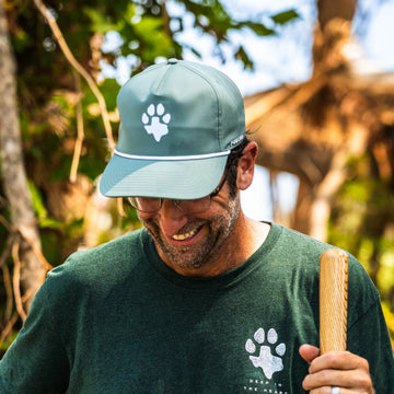 The Deep in the Heart Hat in sage green with a paw print, worn outdoors by a man holding a wooden bat