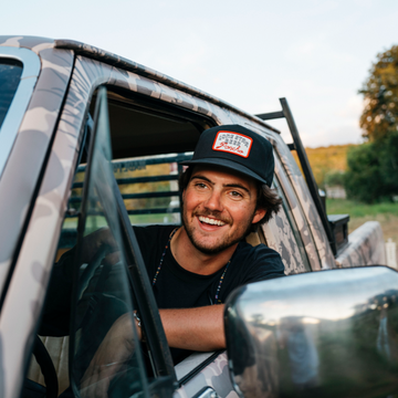 The Dive Hat black cap worn by a smiling man leaning from a camouflaged vehicle outdoors