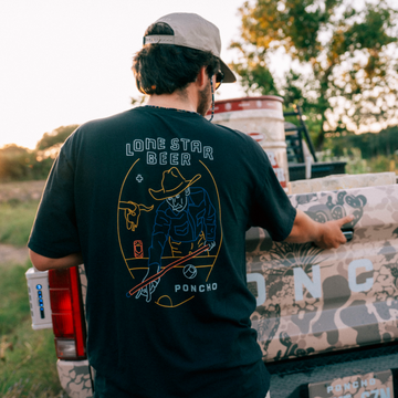 The Dive T-Shirt in black featuring a colorful cowboy graphic, worn outdoors by a pickup truck
